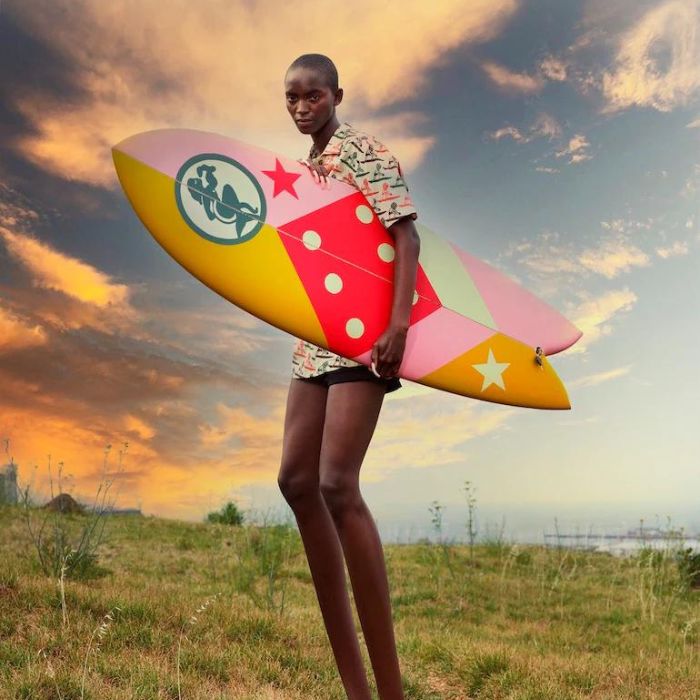 Un jeune homme tient une planche de surf Mami Wata. Il est debout, porte une chemise colorée. Derrière lui, un coucher de soleil