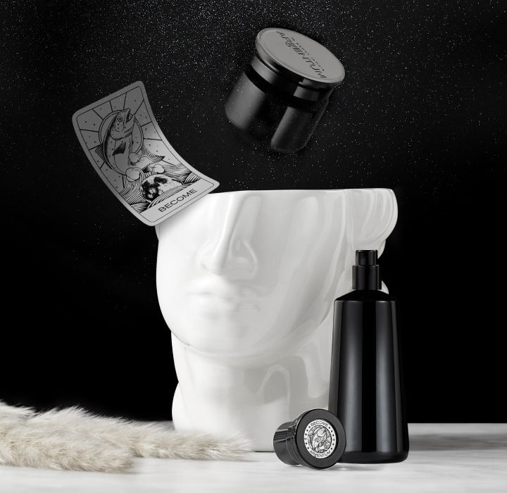 A white sculpted face missing the upper part of its face is placed on a table. Next to it is a bottle of ARgENTUM niche perfume.  A tarot card marked ‘Become’ appears to be flying.