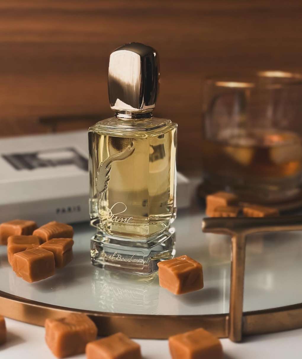 The image shows a perfume bottle from the Electimuss brand, highlighted by colorful lighting. The bottle has an elegant shape, with a red base and shiny gold cap. In the background, colored glass panels in shades of yellow, violet and red create a vibrant, reflective light effect. The luxurious design of the bottle, with a decorative medallion, is accentuated by the reflections of the colors, adding an artistic dimension to the composition.