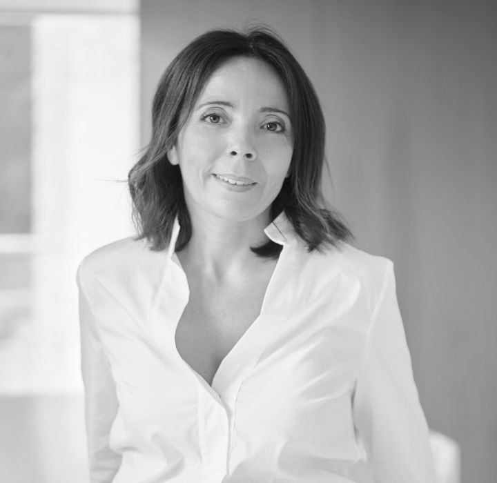 Cette image montre un portrait en noir et blanc d'une femme élégante et souriante, c’est Nadia Benaisa, la fondatrice de la marque de parfums de niche LES SŒURS DE NOÉ. Elle est vêtue d'une chemise blanche classique légèrement ouverte, dégageant une allure professionnelle et raffinée. Ses cheveux sont mi-longs, légèrement ondulés, encadrant son visage.  L’arrière-plan est flou, mettant en valeur le sujet et suggérant un environnement lumineux et moderne. L'ensemble de la photo dégage une impression de sérénité et de confiance.