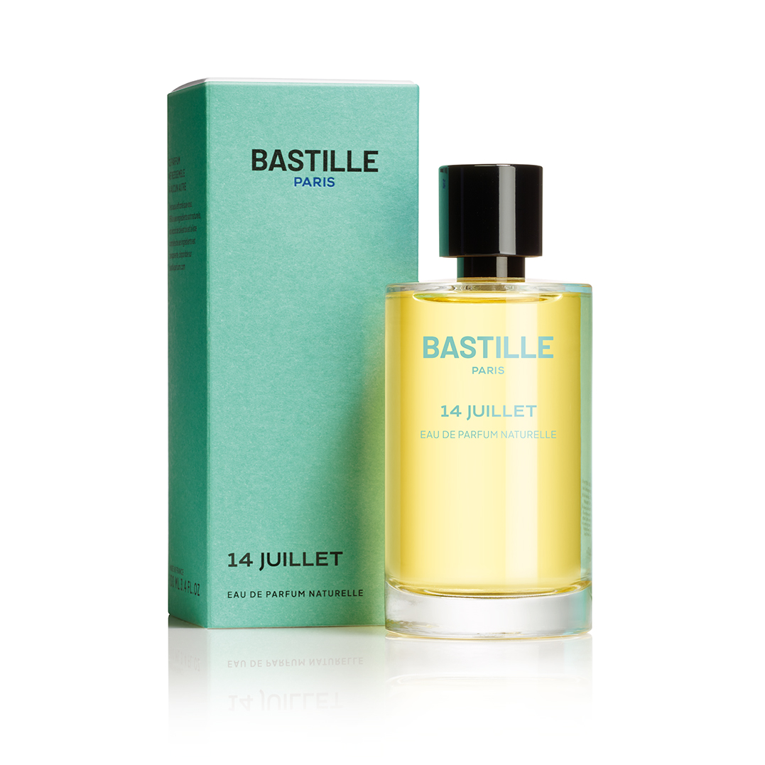 L'image montre un flacon de parfum de la marque Bastille, intitulé "14 Juillet". Le flacon est transparent, laissant voir un liquide de couleur jaune. Il est doté d'un bouchon noir. À côté du flacon, on voit une boîte d'emballage de couleur bleu-vert, avec le nom de la marque et le titre du parfum imprimés en lettres blanches. L'ensemble a une apparence élégante et moderne.