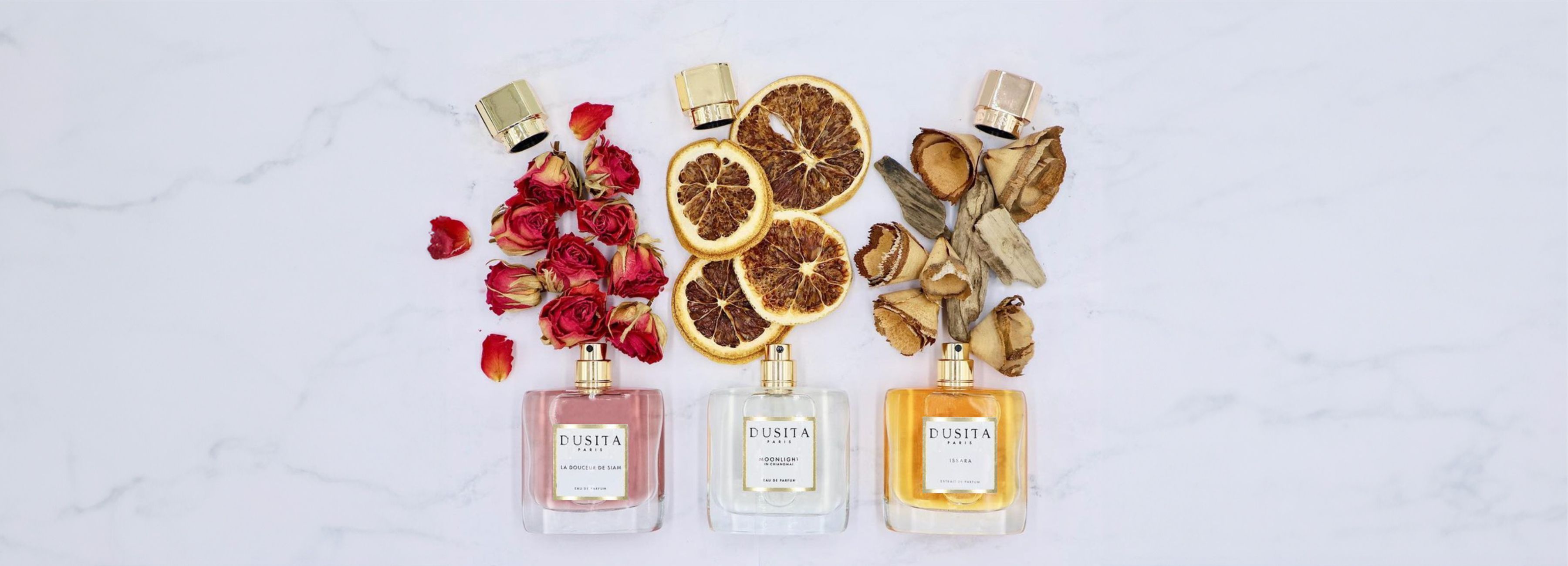 3 flacons de parfums DUSITA posés à plat sur une surface de marbre. Il semble s'en échapper des pétales de roses, des agrumes et des copeaux de bois précieux - SCENTISSIME