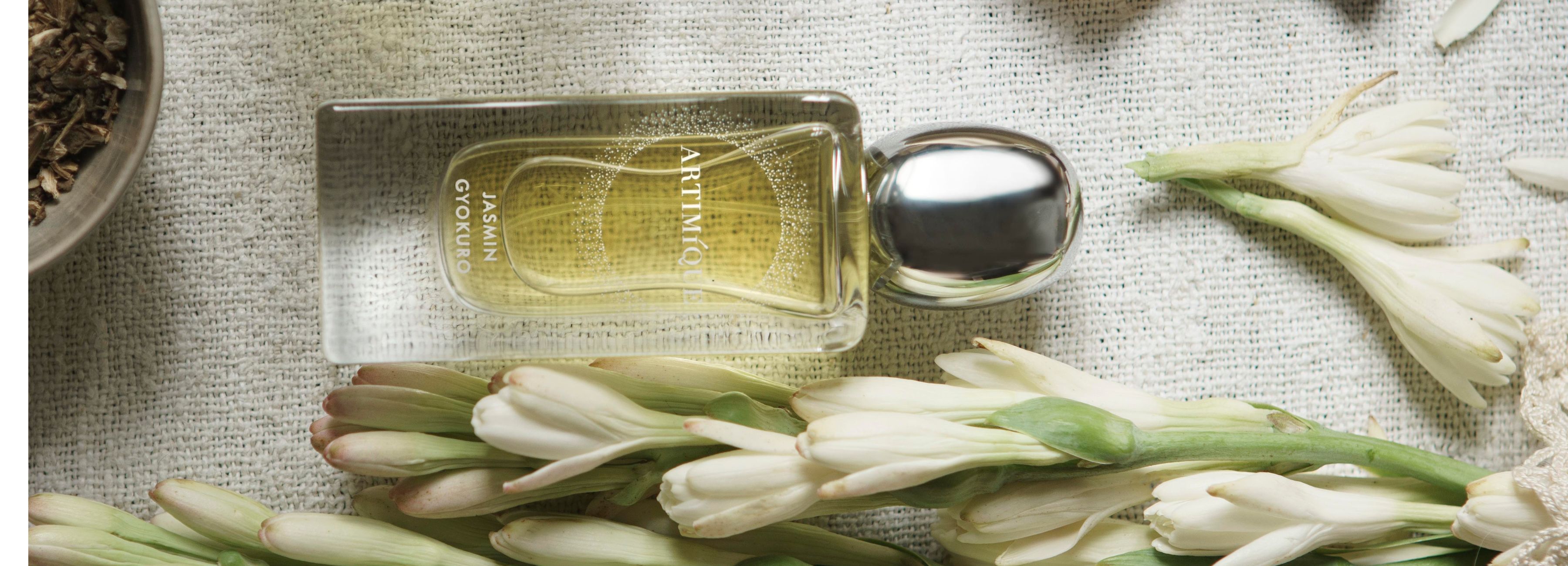 Un flacon de parfum de la marque de niche italienne ARTIMIQUE est posé sur une toile de jute écrue. A côté d'elle on voit des fleurs de jasmin et un bol qui contient des feuilles de thé