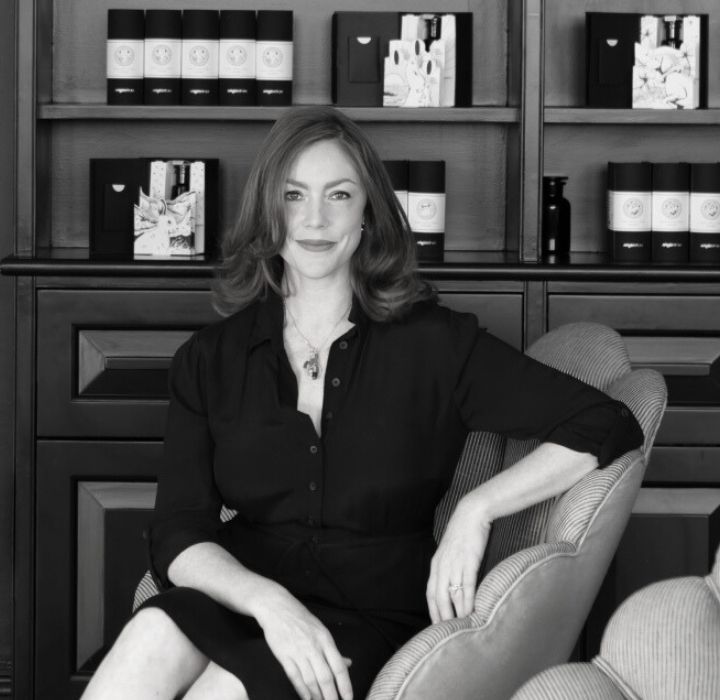 Photograph of Joyce Isaacs, founder of the Argentum brand. She is seated in a leather armchair. Behind her is a bookcase displaying several Argentum products.