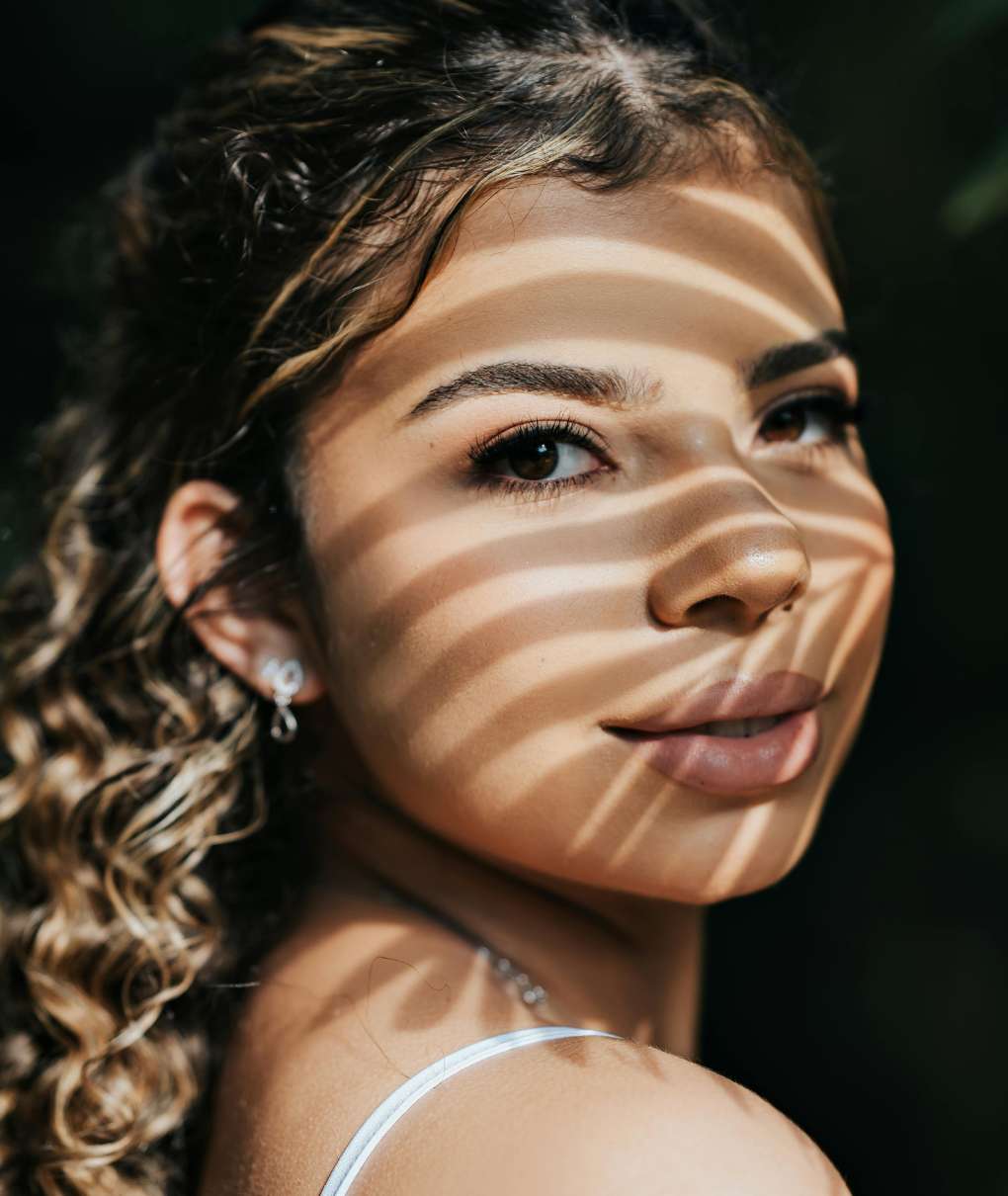 The image is a portrait of a young woman, looking over her shoulder with a slight smile. Her face is partially shaded by horizontal lines of light, as if she were standing in the shadow of a slatted structure or foliage.Her hair is curly and appears to have lighter strands, framing her face. She is wearing a thin garment strap, suggesting a light outfit.The background is dark and out of focus, emphasizing the woman's face and creating a striking contrast with the lit areas.