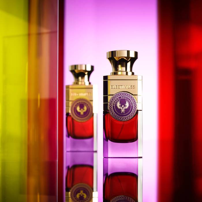 L'image montre un flacon de parfum de la marque Electimuss, mis en valeur par un éclairage coloré. Le flacon a une forme élégante, avec une base rouge et un bouchon doré brillant. À l'arrière-plan, des panneaux de verre colorés dans des tons de jaune, violet et rouge créent un effet de lumière vibrante et réfléchissante. Le design luxueux du flacon, avec un médaillon décoratif, est accentué par les reflets des couleurs, ajoutant une dimension artistique à la composition.