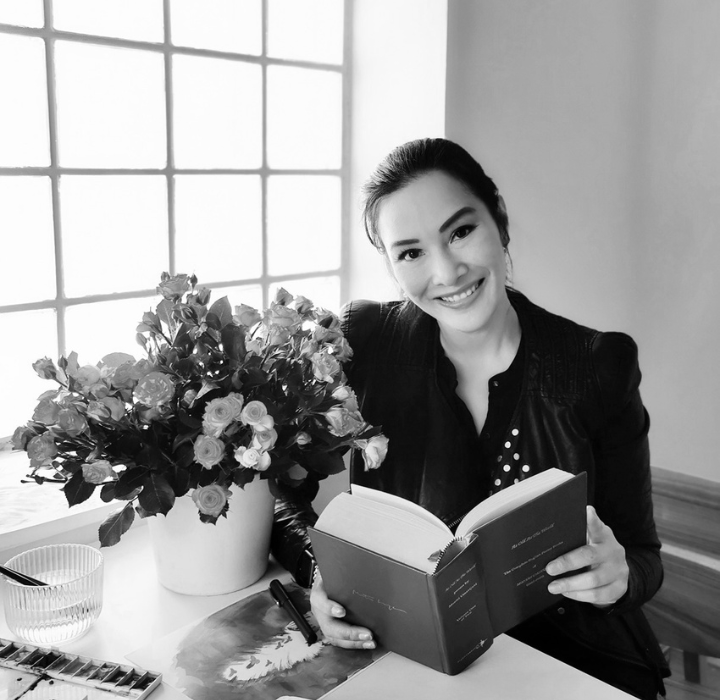 Portrait de Pissara Umajivani. Elle est assise à un bureau sur lequel il y a un bouquet de fleurs. Elle tient une gros livre ouvert. Elle est souriante.