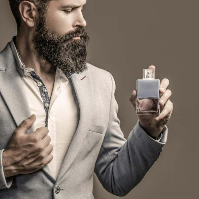 L'image montre un homme élégant, portant un costume gris clair. Il a une barbe bien entretenue et regarde avec attention un flacon de parfum qu'il tient dans sa main. Son expression est réfléchie, et l'éclairage met en valeur les détails de son visage et de sa tenue. Le fond est neutre, ce qui accentue la sophistication et le style du personnage.