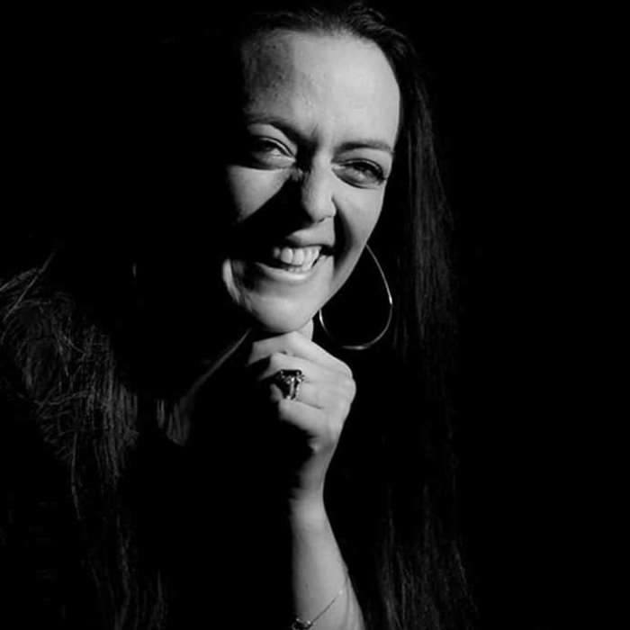 De afbeelding toont een glimlachende vrouw in donkere verlichting. Haar gezicht is gedeeltelijk verlicht, waardoor haar stralende glimlach en sprankelende ogen worden benadrukt. Ze heeft lang haar dat vrij rond haar gezicht valt.Ze draagt grote, opvallende oorbellen en een zichtbare armband om haar pols. Haar hand is opgeheven dicht bij haar gezicht in een vriendelijk gebaar.